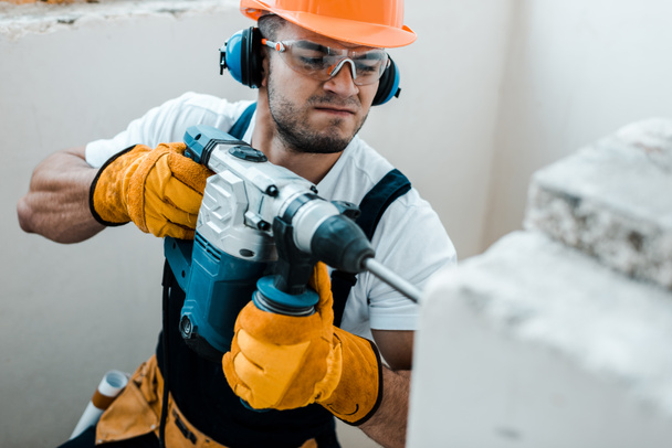 selective focus of handsome handyman in uniform and yellow gloves using hammer drill  - Photo, Image