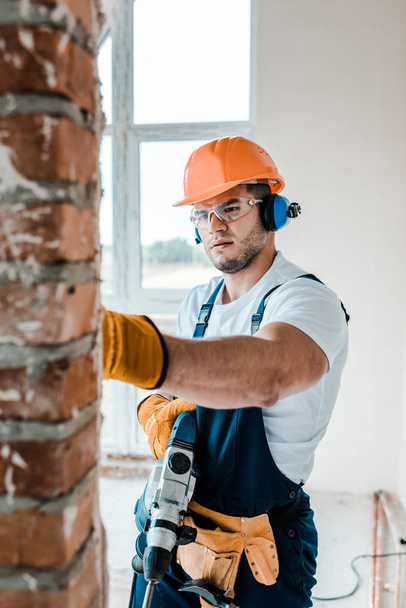 selective focus of workman in uniform and yellow gloves holding hammer drill and looking at brick wall  - Foto, imagen