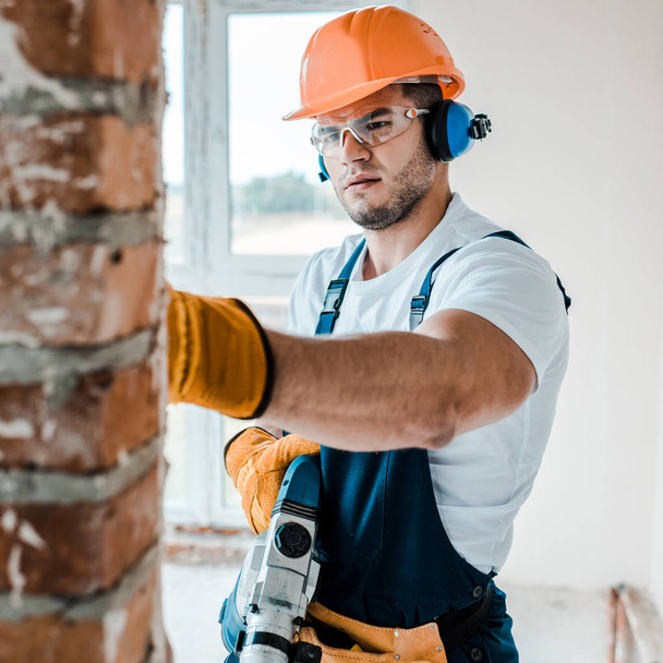 selective focus of workman in uniform and goggles  holding hammer drill and looking at brick wall  - Photo, Image