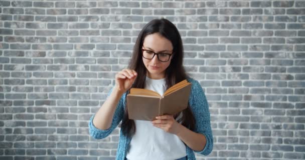Serious girl in glasses reading book turning page on brick wall background - Materiał filmowy, wideo