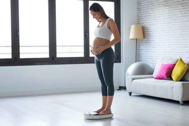 Pretty young pregnant woman standing on scales at home. - Photo, Image