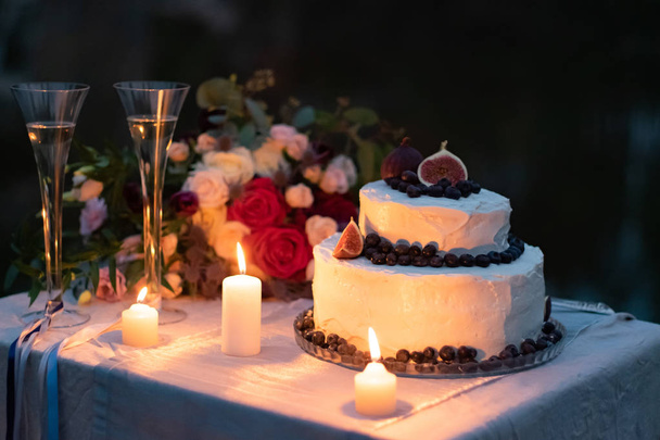 svatební dekorace. dort v bílé Ledovka s dekoracemi borůvek a fíků na stole, večer s brýlemi, zapálenou svíčkami a svatební kyticí - Fotografie, Obrázek