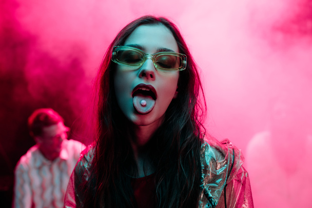 beautiful girl in sunglasses with lsd on tongue in nightclub with pink smoke - Photo, Image
