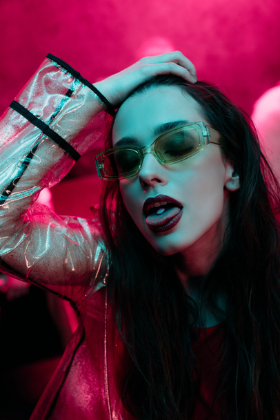 ελκυστικό κορίτσι σε γυαλιά ηλίου με LSD στη γλώσσα σε νυχτερινό κέντρο με ροζ καπνό - Φωτογραφία, εικόνα