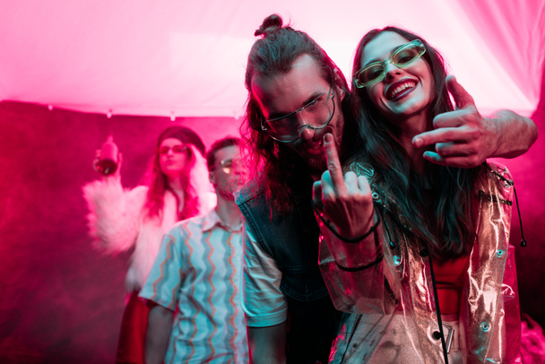 ευτυχισμένο κορίτσι και ο άνθρωπος σε γυαλιά ηλίου που δείχνουν μεσαία δάχτυλα στο νυχτερινό κλαμπ κατά τη διάρκεια ρέιβ πάρτι  - Φωτογραφία, εικόνα