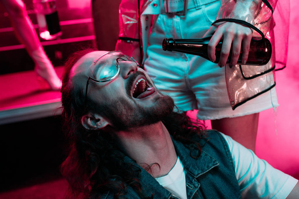 woman pouring alcohol in mouth of man in sunglasses with lsd on tongue in nightclub  - Foto, Imagen