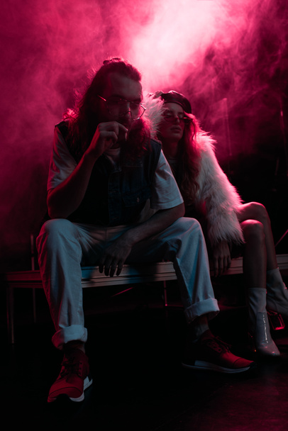 man smoking cigarette near young woman during rave party in nightclub with pink smoke - 写真・画像