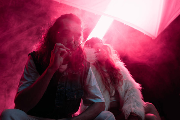 man smoking cigarette near young woman during rave party in nightclub - 写真・画像