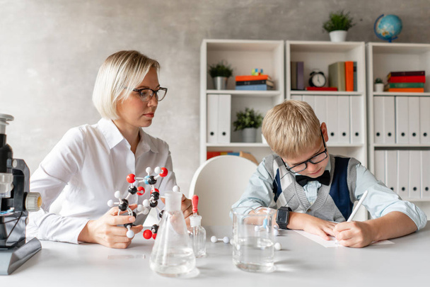 Blond schoolboy write properties of chemical element into his notebook under guidance of his charming teacher - Photo, image