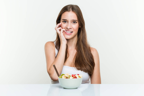 Young caucasian woman eating fruit bowl biting fingernails, nervous and very anxious. - Photo, Image