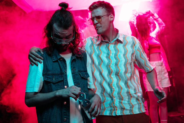 man in zonnebril knuffelen een andere man met waterfles in nachtclub tijdens rave party  - Foto, afbeelding