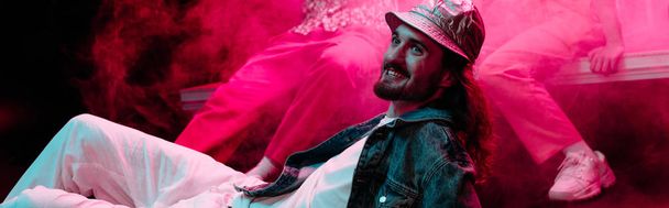 panoramiczny strzał uśmiechniętego człowieka leżącego na podłodze w klubie nocnym podczas rave party z różowym dymem - Zdjęcie, obraz