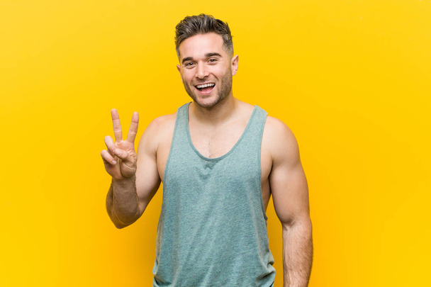 Young fitness man against a yellow background showing victory sign and smiling broadly. - Photo, Image