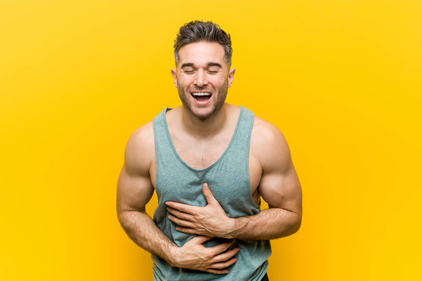 Joven fitness hombre contra un fondo amarillo se ríe felizmente y se divierte manteniendo las manos en el estómago
. - Foto, Imagen