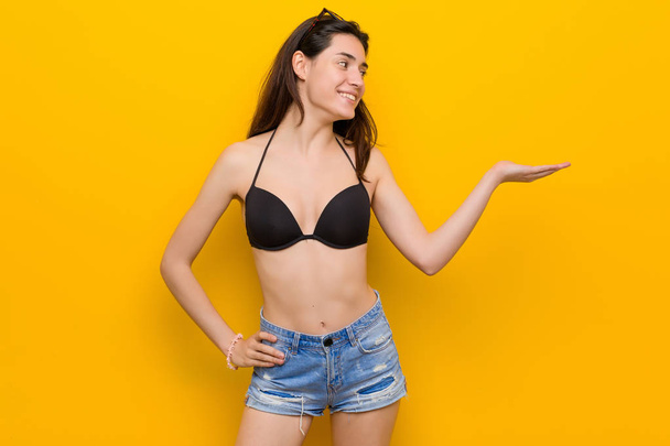 Nuori brunette nainen yllään bikinit vastaan keltainen tausta osoittaa kopio tilaa kämmenellä ja tilalla toinen käsi vyötäröllä
. - Valokuva, kuva