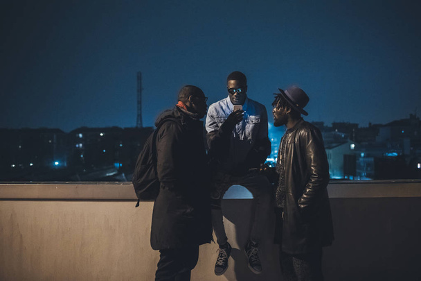Üç Afrikalı arkadaş dışarıda oturuyor ve akıllı telefonlara bakıyor. - Fotoğraf, Görsel