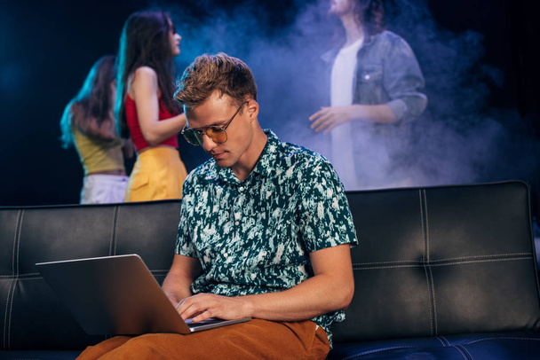 homme en lunettes de soleil assis sur le canapé et en utilisant un ordinateur portable dans la boîte de nuit
 - Photo, image