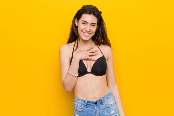 Young brunette woman wearing a bikini against yellow background laughs out loudly keeping hand on chest. - Foto, Imagen