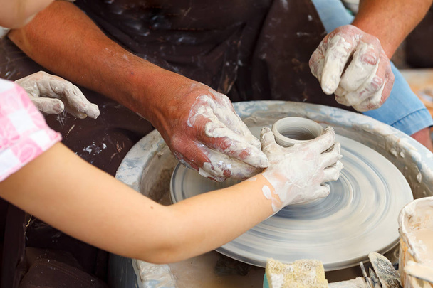 陶器をクローズアップ。車輪の上の粘土を助けるために子供の手を導く大人の陶芸家泥だらけの手 - 写真・画像