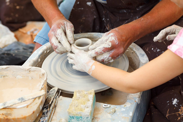 Закрыть керамику. Взрослые грязные руки горшечника, направляющие детские руки, чтобы помочь с глиной на колесе
 - Фото, изображение