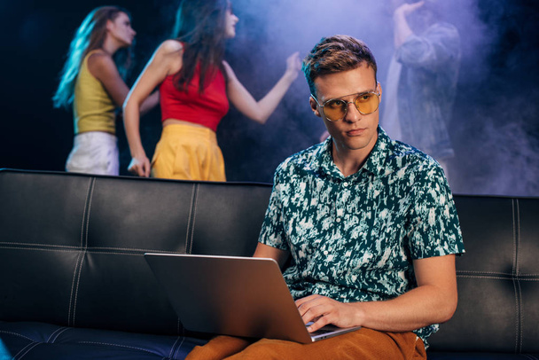 man in sunglasses sitting on couch and using laptop in nightclub - Photo, image