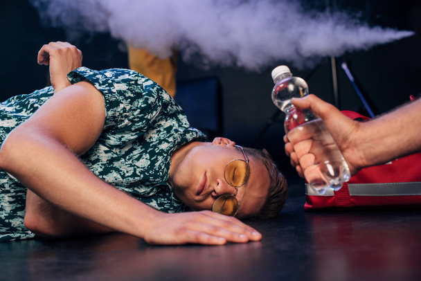 человек в солнцезащитных очках лежит на полу возле бутылки воды в ночном клубе
 - Фото, изображение