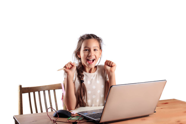 A 10 years old school girl sitting at a table with a laptop with an expressive emotional face isolated on white. Successful pass of exam, good scores for test, online education, homeschooling concept - Photo, image