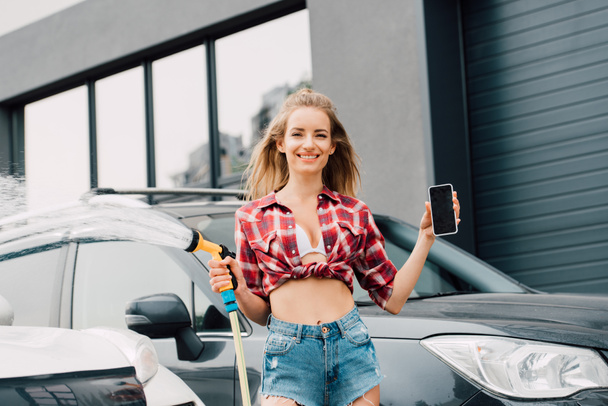cheerful girl holding smartphone with blank screen and portable washer near cars  - Photo, Image