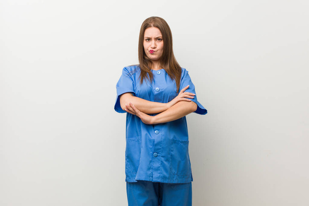 Νεαρή νοσοκόμα γυναίκα εναντίον ενός λευκού τοίχου Συνοφρυωμένο πρόσωπο σε δυσαρέσκεια, κρατά τα χέρια διπλωμένα. - Φωτογραφία, εικόνα