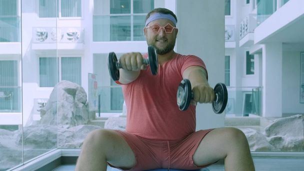Funny fat male in pink glasses and in a pink t-shirt is engaged with dumbbells on a fit ball in the gym depicting a girl - Φωτογραφία, εικόνα