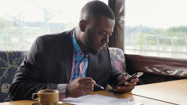 Ο Αφροαμερικανός επιχειρηματίας γεμίζει έγγραφα σε θερινό καφέ σκηνής κοιτάζοντας στο smartphone. - Φωτογραφία, εικόνα