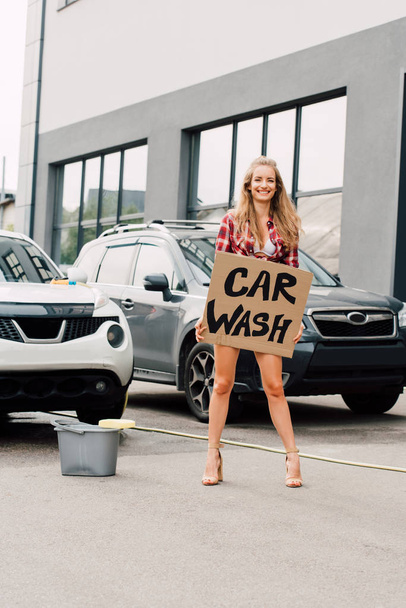 positive girl standing and holding carton board with car wash lettering near cars  - Foto, afbeelding