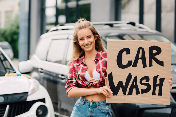 happy girl holding carton board with car wash lettering near cars  - Foto, afbeelding