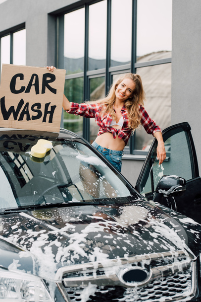 cheerful young woman holding carton board with car wash lettering and standing near automobile  - Photo, Image