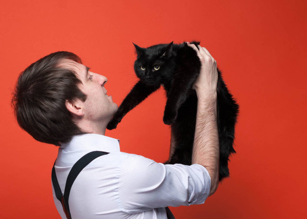 side view of handsome man in shirt with rolled up sleeves holding over face cute black cat, smiling and looking at it muzzle in front of coral background with copy space - Foto, Bild