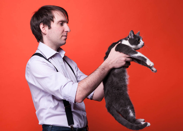 side view of handsome man in shirt with rolled sleeves holding cute gray and white cat and looking at it back in front of orange background with copy space - Foto, immagini