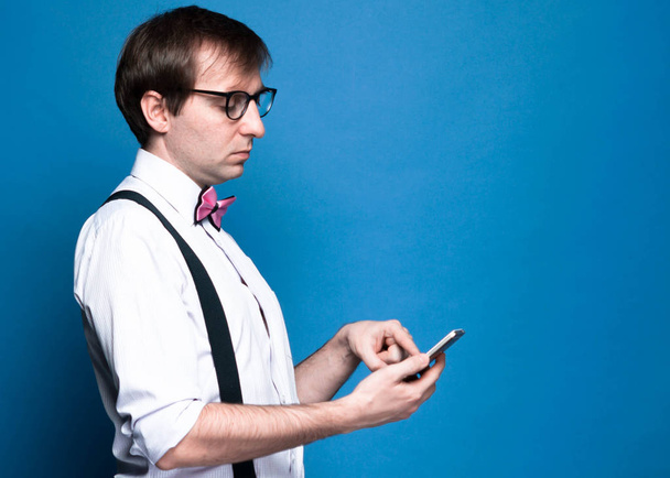side view of handsome man with dark hair in pink shirt with rolled up sleeves, bow tie, black suspender and glasses using smartphone on blue background with copy space - Foto, Imagen
