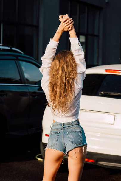 πίσω όψη της νεαρής γυναίκας με σορτς στέκεται κοντά σε σύγχρονα αυτοκίνητα  - Φωτογραφία, εικόνα