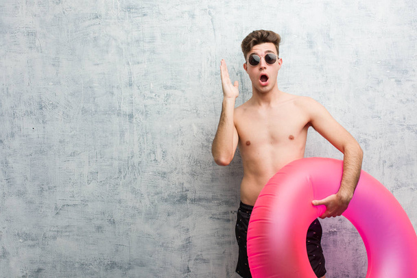 Young man holding a pink inflatable donut wearing a swimsuit surprised and shocked. - Photo, Image