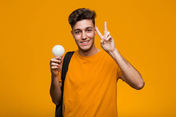 Young student man holding a light bulb looking sideways with doubtful and skeptical expression. - Photo, Image
