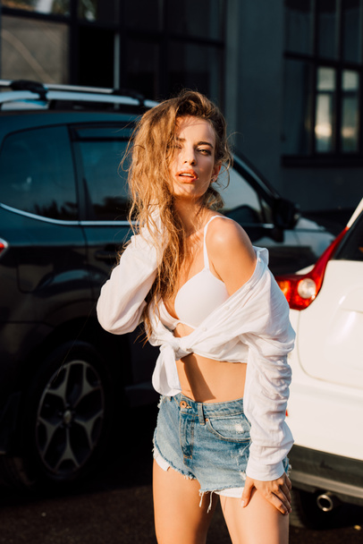 attractive woman in blue shorts looking at camera and touching hair near cars  - Foto, immagini