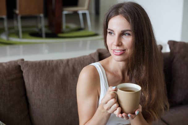 portrait of beautiful smiling brunette girl sitting on sofa in living room and holding mug of tea in her hands. Girl looks away - Foto, Imagen