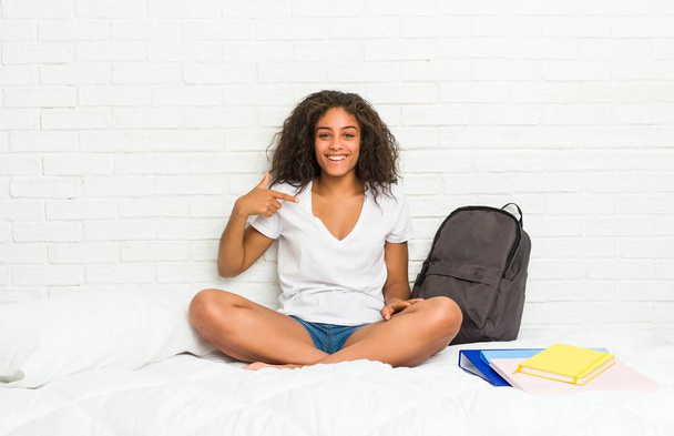 junge afrikanisch-amerikanische Studentin auf dem Bett Person zeigt mit der Hand auf ein Hemd Kopierraum, stolz und zuversichtlich - Foto, Bild