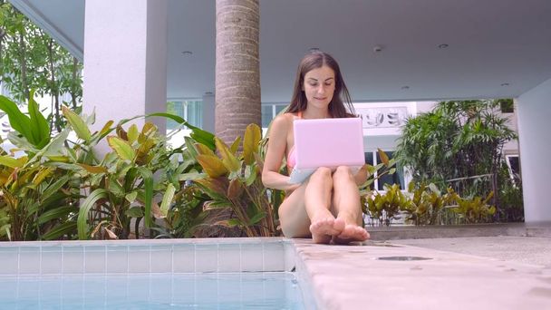 Mladá ženská na volné noze, která sedí s notebookem poblíž bazénu. Pohled zpět. Během svátků je obsazeno. Koncept vzdálené práce. selektivní zaměření - Fotografie, Obrázek