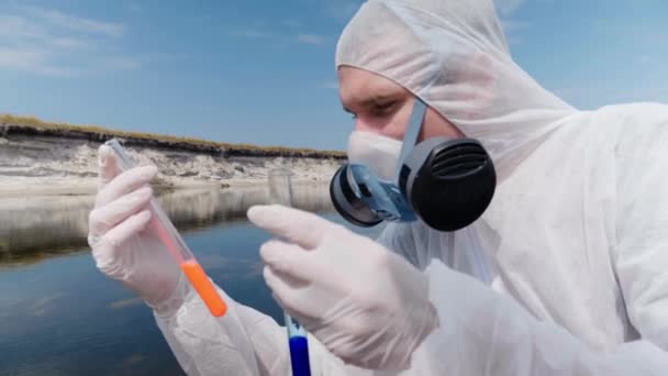 Man in a protective suit and respirator observes a chemical reaction of water in test tube, which he took for analysis from polluted river. Scientist takes samples in vitro. Ecological disaster. 4K - Footage, Video
