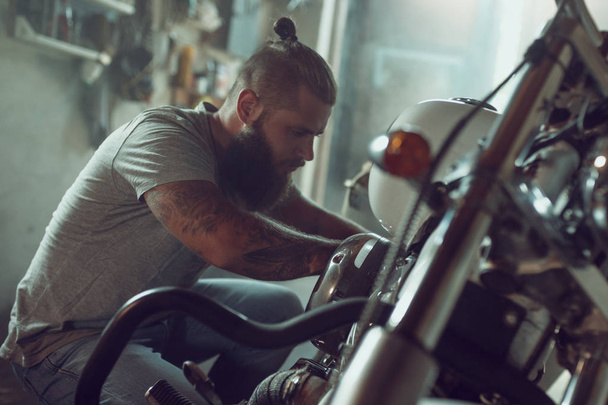 Όμορφος άντρας με γένια που επισκευάζει τη μοτοσικλέτα του στο γκαράζ. Ένας άντρας που φοράει τζιν και ένα μπλουζάκι - Φωτογραφία, εικόνα