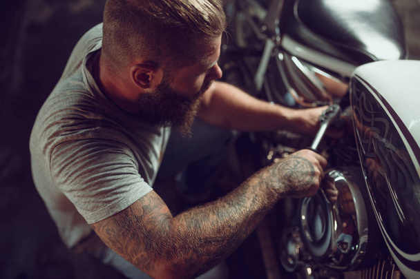 レザー ジャケットと太陽メガネでハンサムなひげを生やした男は修理店のバイクに座っています。 - 写真・画像