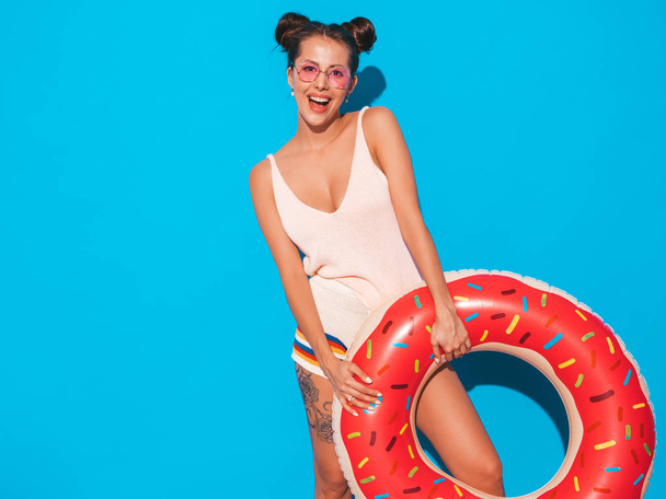Young beautiful sexy smiling hipster woman in sunglasses.Girl in summer clothes with donut lilo inflatable mattress.Positive female going crazy.Funny model isolated on blue - Foto, afbeelding