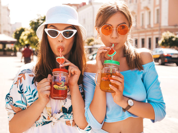 Two young beautiful smiling hipster girls in trendy summer clothes and panama hat.Sexy carefree women posing in the street.Positive models having fun in sunglasses.Drinking fresh cocktail smoozy drink - Φωτογραφία, εικόνα