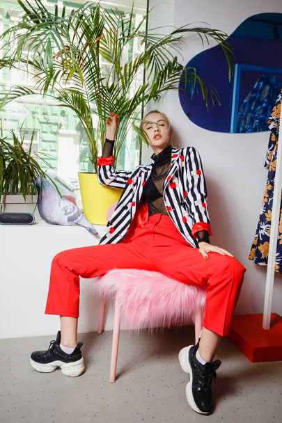 Fashion young girl blogger dressed in stylish striped jacket and red trousers poses sitting on the stool with pink fur in the room on the background with plant - Foto, Imagem
